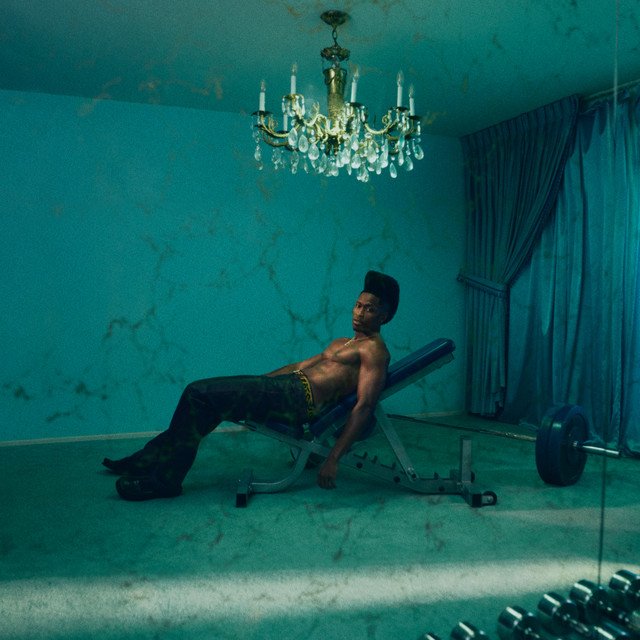 ALOUD! album cover: Lucky Daye shirtless, leaning back on a weight bench in an empty teal room with a crystal chandelier